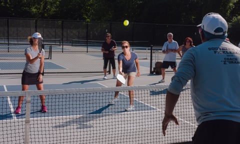 A man stands on one side of a low net on a pickleball court facing two women, all holding paddles.