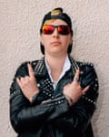 A woman with short yellow blond hair in a black silver studded leather jacket and a white shirt wearing red reflective shades and a back to front peaked cap with her arms crossing her chest