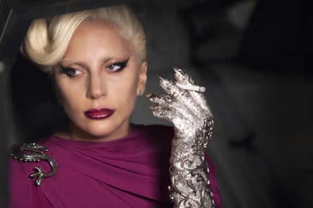 Head and shoulders of a glamorous young woman with side parted 1940s style peroxide blond hair wearing a purple neck high dress a serpent brooch clipped to the left and smoking a cigarette in one long silver gloved hand