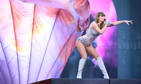 Taylor Swift performs at the Melbourne cricket ground: she is wearing a sparkly leotard-type garment with knee-length silver high-heeled boots, and is singing in front of a pink-lit backdrop