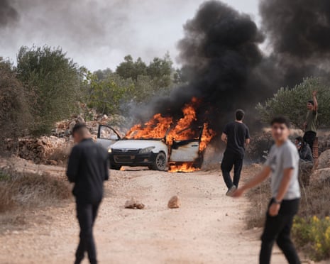 A car on fire in the middle of a dirt track with people stood around.