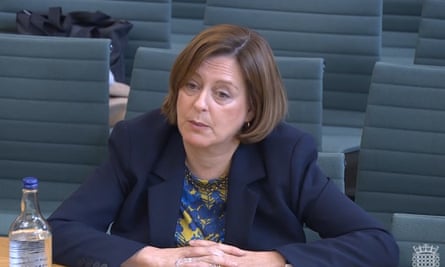 Melanie Dawes, seen here in the House of Commons addressing a select committee: she is in her late 50s and has brown chin-length, straight hair, and wears a dark blue jacket over a yellow and blue patterned blouse; she is leaning on a desk with her hands clasped in front of her