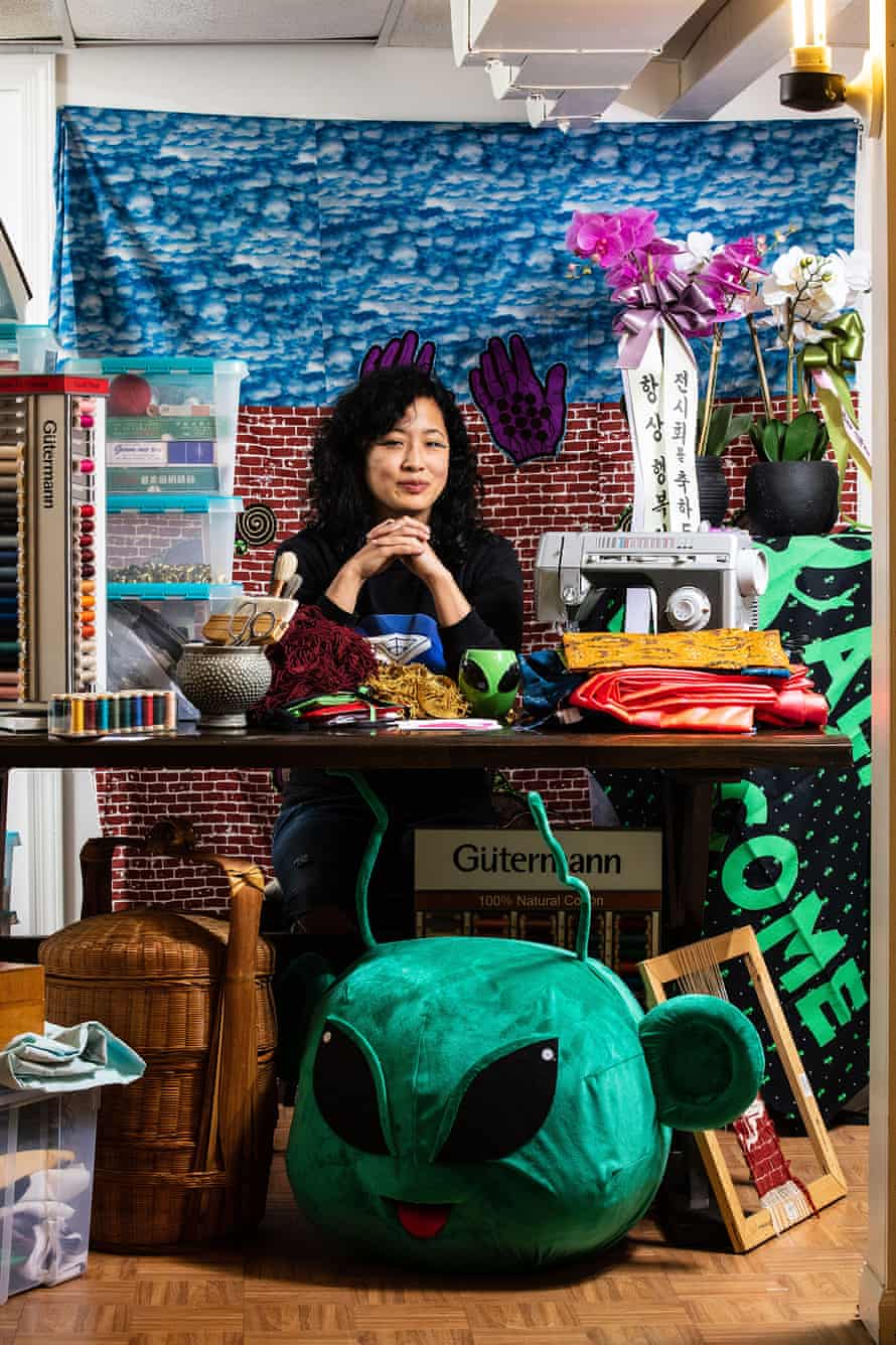 A woman sits at a desk, surrounded by fabrics and other textiles. At her feet under the desk is a large green alien head made out of fabric.
