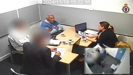 Low angle CCTV image of Ryland Headley with crossed arms in the corner seat of an interrogation room with four officers facing him