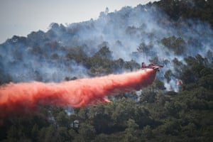 A light aircraft flies low over a hillside of smouldering trees leaving a trail of red powder behind it
