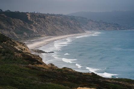 A view of an oceanside cover, with a thin sandy beach between frothy ocean and vegetation-covered cliffs.