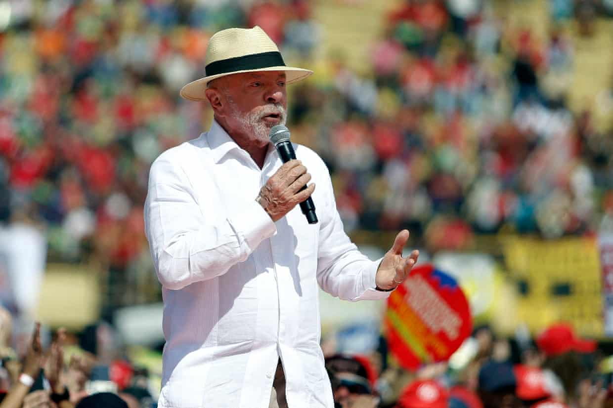 President Lula, wearing a panama hat and white shirt, speaks into a microphone, surrounded by a huge crowd