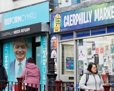 People walking past a shop front with a large photo of Nigel Farage on it, next to a convenience store