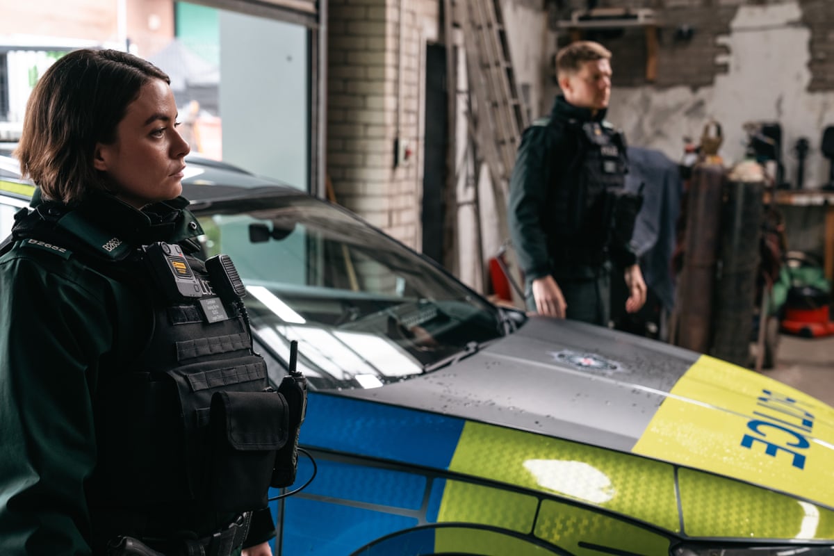 The two actors, playing police officers, stand in uniform either side of a police car