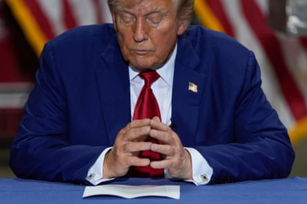man in blue suit and red tie sits at a blue table with fingertips pressed together