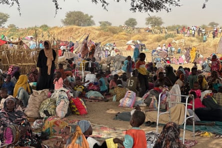 A crowd of African women and children sit in the shade of a tree with their belongings. Two walking frames can be seen among the bundles of possessions