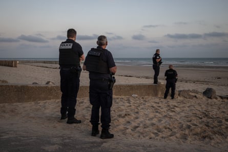 Four police officers in black uniform stand on the beach, one on a stone breakwater, looking out to sea. There are small clouds in the sky but the sea looks calm.