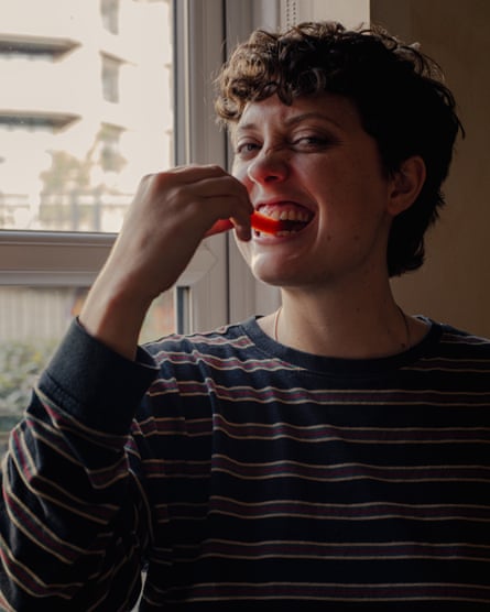 Lucy Knight smiles widely while biting into a slice of red pepper near a window