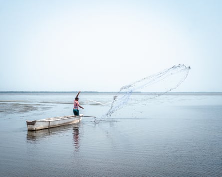 A man stands on a small boat throwing a fishing net out over calm water