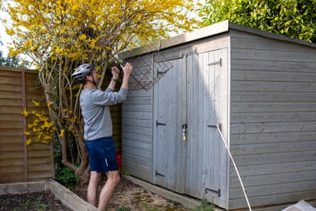 Man wearing a cycling helmet holding a large cage with both hands, standing in front of a garden shed.