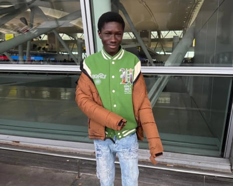Javits Frimpong, smiling, in a green and brown jacket, stands in an airport terminal