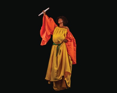 A woman stands in a statuesque pose draped in a yellow dress and orange cloak with a raised fist holding a scroll
