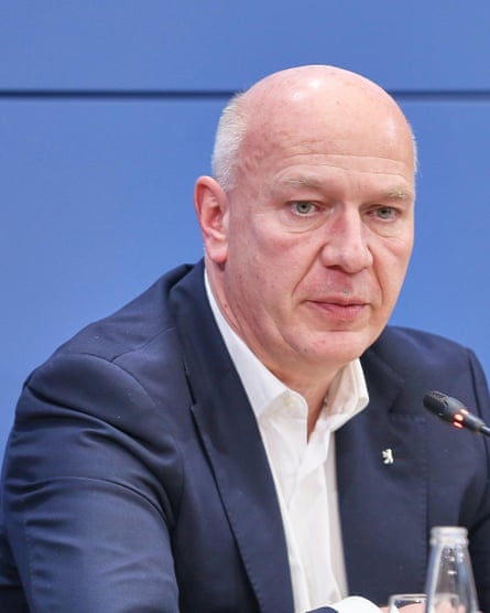 he is in his early 50s and near-bald with white hair, and wears a dark blue jacket and white shirt. He looks downward and is unsmiling.