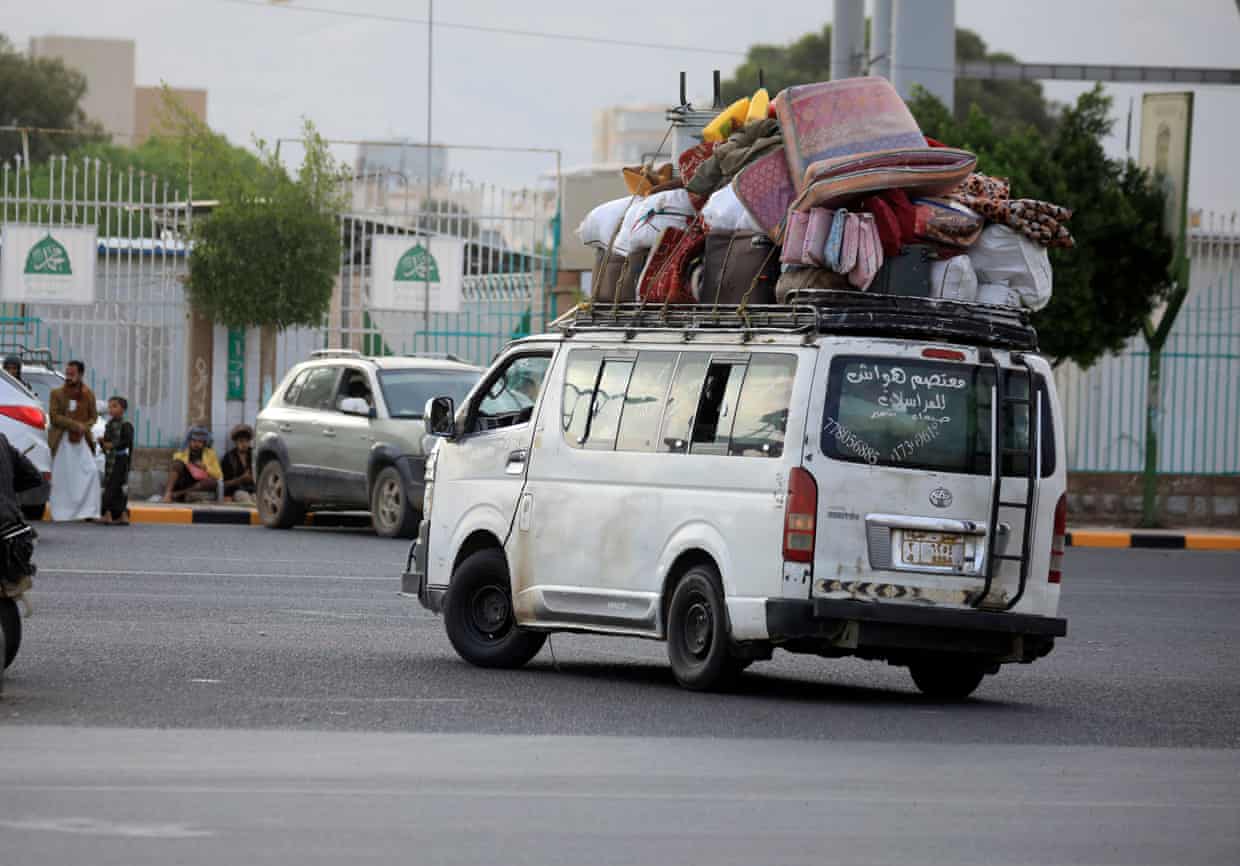 A white minibus on a street has mattresses, cushions, bedding and other baggage strapped to its roof.