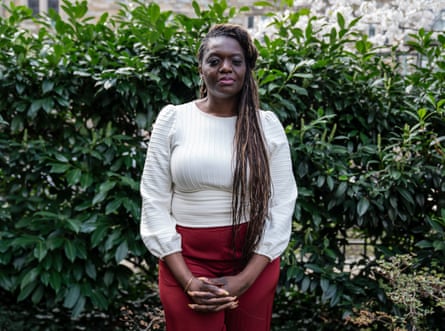 A Black woman with long dark brown braids wearing a white blouse and red pants stands in front of a shrub with green leaves.