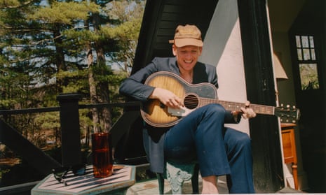 Willie J Healey smiling, playing his guitar, seated on the porch of a house in the woods