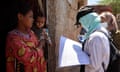 A health worker in protective gear, carrying a large checklist, approaches a mother and baby at their door