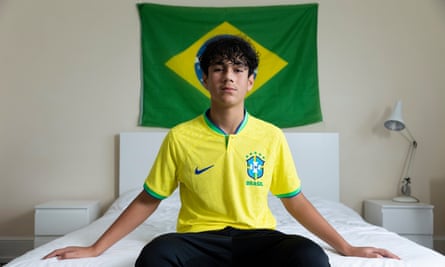 Nicholas Goncalves-Newman Brazil sitting on a bed wearing a yellow-and-green Brazil shirt and with a Brazilian flag on the wall behind him