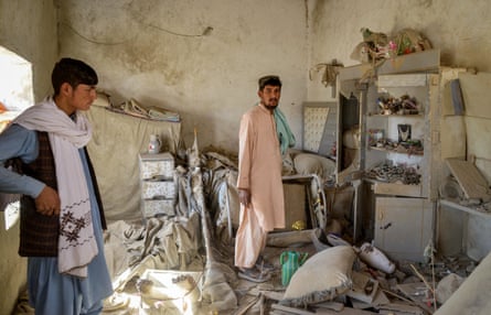 a young man with short dark hair and an older man with a beard stand in a ruined room with debris on the floor and curtains and household items strewn around. Both wear long robes and scarves.