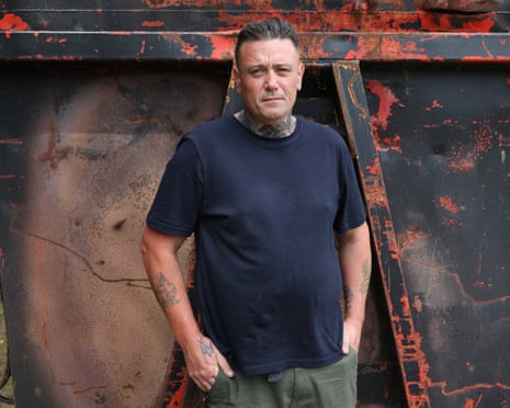 Micky Steeds, with tattoos and slicked-back hair, stands in a dark t-shirt against a red and dark backdrop