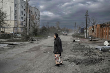 An elderly woman looks sadly down a street full of damaged buildings under a bleak grey sky