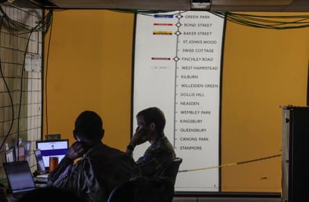 Soldiers sit in front of laptops on the London Underground platform with a train route in the background