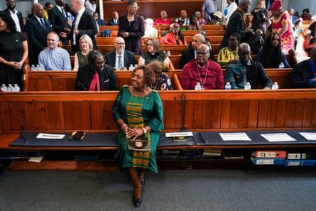 Olivia Grange sits smiling in a front-seat pew with lines of people, mostly black but also a few white, behind her including several in clerical collars. She is 80 years old with short curly hair worn in ringlets and is wearing a dark green dress with gold embroidery.