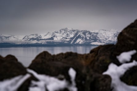 Blurred snowy rocks successful nan foreground, pinch a reservoir beyond, and snowy mountains connected nan different side.