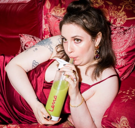 Lena Dunham in red dress, leaning against red cushions, drinking green liquid out of a water bottle