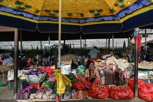 A woman sits in the foreground under a parasol in a market, with vegetables laid out around her