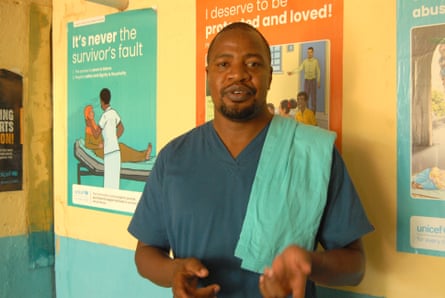 A man in blue scrubs stands against a wall covered in posters bearing health messages.