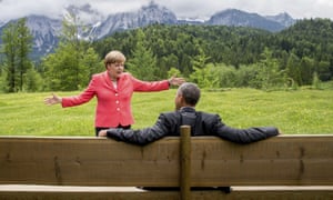 The back of Barack Obama sitting on a bench with his arms spread on the back of it and German Chancellor Angela Merkel in a pink jacket facing him with her arms spread, and a grassland and snowy mountains behind