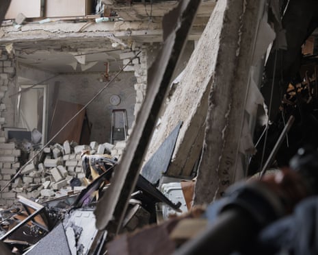 A destroyed apartment interior with rubble scattered across the floor, collapsed ceiling beams, and exposed walls visible through a metal frame in the foreground, following a drone strike on a multi-story residential building in Kharkiv