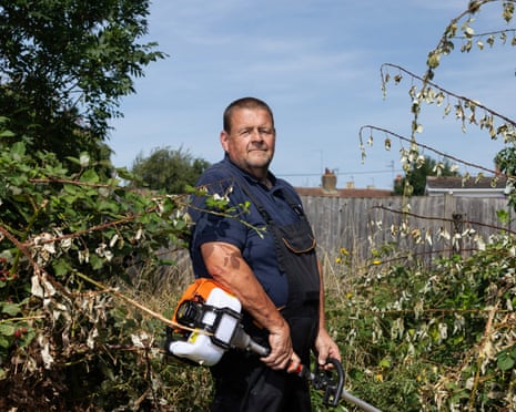 Wearing black dungarees over a blue polo shirt, he holds a garden trimmer amid an overgrown garden and poses for the camera