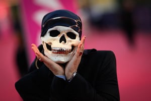 A man in a black suit and a skull mask with silver teeth cups his hands under his chin
