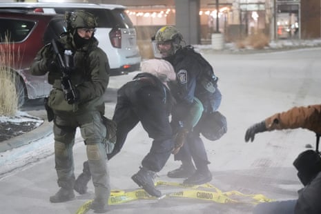 A law enforcement officers in riot gear is pulling a person in full winter gear face-first onto the ground while another law enforfement officer holds a weapon pointed to the ground as he watches a group of other protesters.