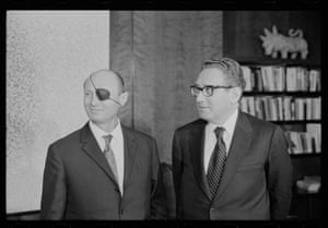 A man with a black eye patch stands with Kissinger in front of bookshelves