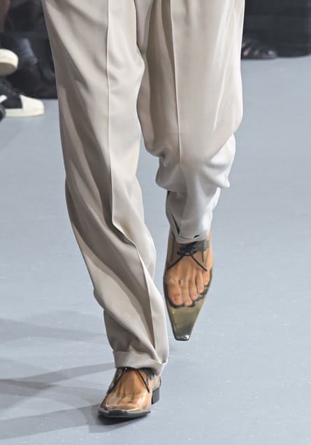 A male model’s legs walk down a runway with see-through plastic shoes on the feet