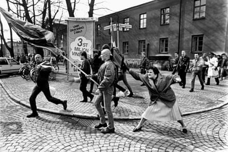 A woman moves up behind a flag-carrying skinhead and swings her handbag at his head