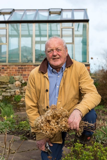 A man in a tan coat holds a dried bushel near a greenhouse.