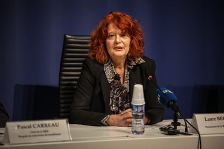 she sits at a long table behind a microphone and wears a black jacket and black blouse with white pattern; she is in her mid-60s and has bright red hair.