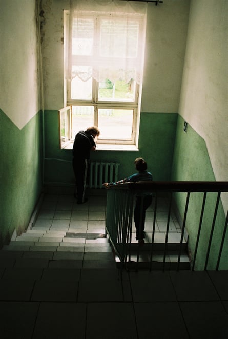 Two children stand at a window on the landing of a tiled staircase.