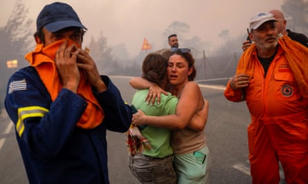 Two women embrace on a road as men in overalls and uniforms look towards a fire