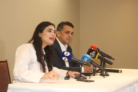 A Latina woman speaking at a table covered in microphones with a man next to her