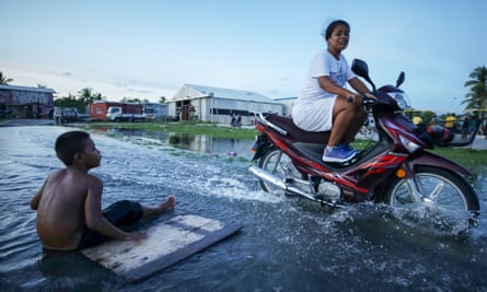 A woman on a motorbike rides through flood water past a boy apparently floating on a piece of wood